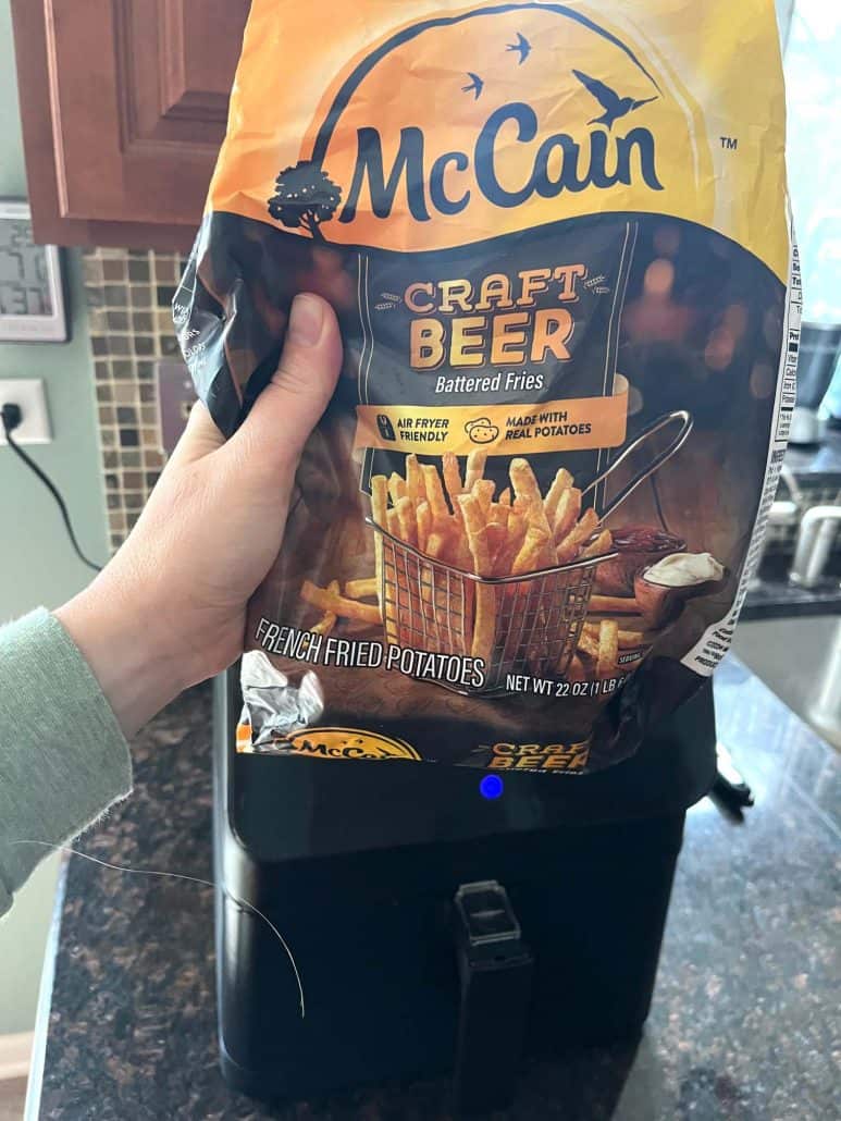 Hand of Melanie Mendelson from melaniecooks.com holding a bag of McCain Craft Beer Battered Fries over an air fryer, ready to cook.